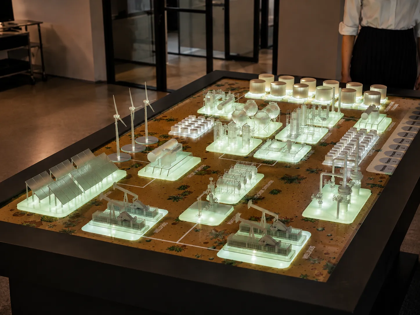 Illuminated interactive touch-table model of an energy facility with wind turbines and refinery units, by The Monolith
